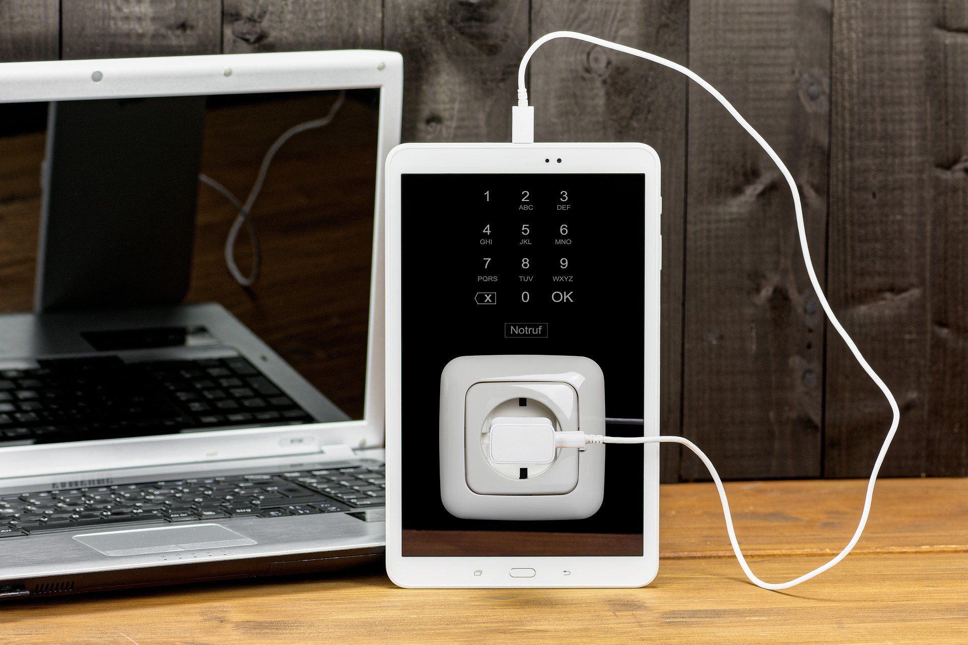Ein weißes Tablet, das über USB mit einem Laptop verbunden ist, zeigt einen Tastaturbildschirm und ein Bild von Smarte Steckdosen mit eingestecktem Ladegerät. Beide Geräte stehen auf einer hölzernen Oberfläche vor einer dunklen Holzwand im Hintergrund.