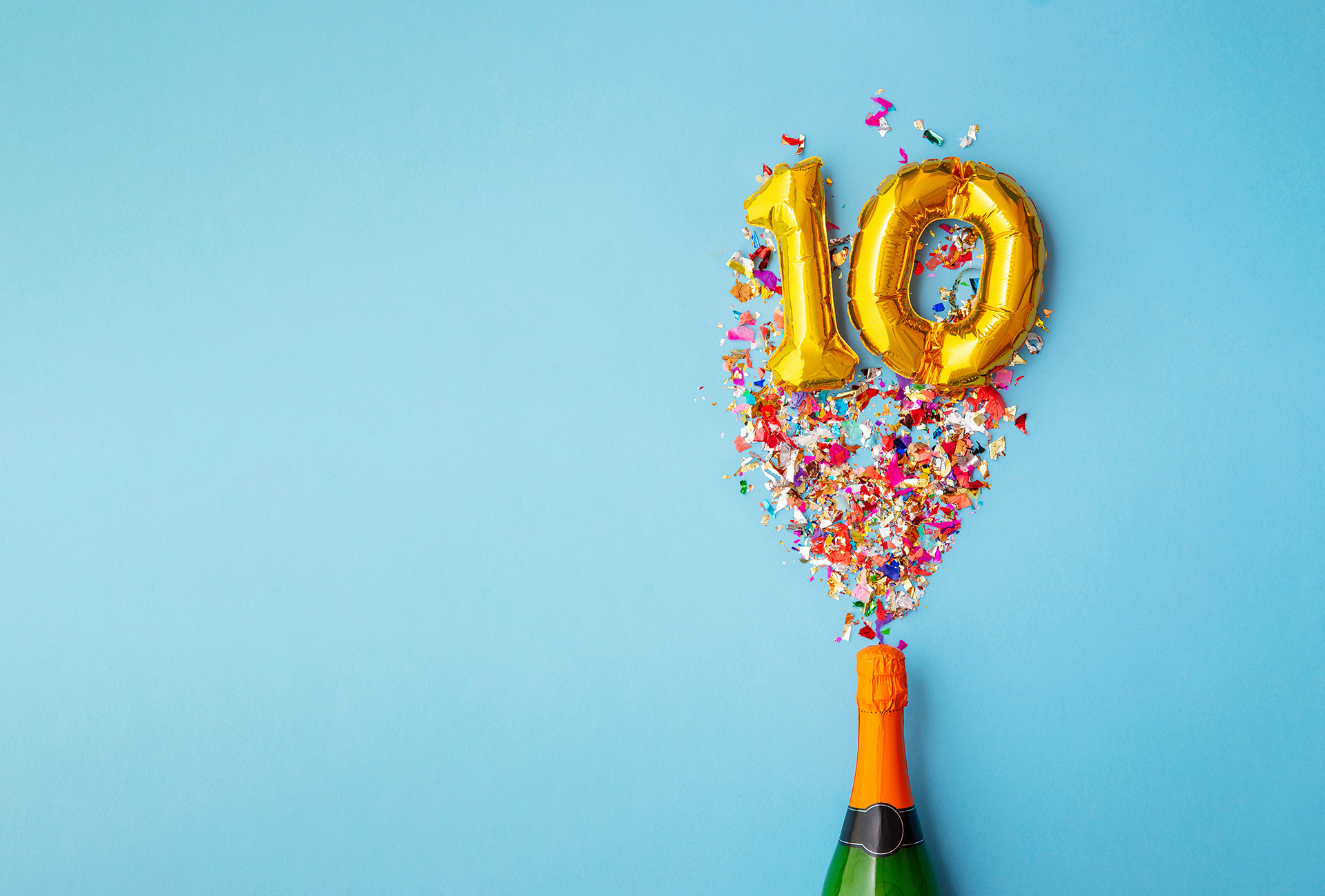 Eine Sektflasche knallt auf, gibt buntes Konfetti und goldene Luftballonnummern "1" und "0" vor blauem Hintergrund frei und feiert ein 10-jähriges Jubiläum oder Firmenjubiläum in festlichem Stil.