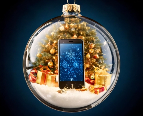 Ein gläserner Weihnachtsschmuck enthält einen geschmückten Baum, eingepackte Geschenke, Kunstschnee und ein Smartphone mit blauem, verschneitem Bildschirm - alles vor einem dunkelblauen Hintergrund, der uns daran erinnert, in der Weihnachtszeit auf Phishing zu achten.
