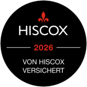 Schwarzes kreisförmiges Abzeichen mit einer roten Lilienblüte oben, dem Wort HISCOX in Weiß, 2026 in Rot und VON HISCOX VERSICHERT in Weiß, das als Versicherungsimpressum von Hiscox dient.