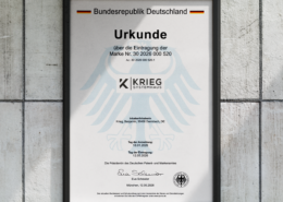 Eine gerahmte deutsche Urkunde mit dem Titel „Urkunde“, die ein schwarzes Adlerwappen und das Logo von KRIEG SYSTEMHAUS trägt und an einer hellen Wand hängt.